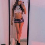 Petite mixed race Chaturbate model Alex_Saeli wearing a short metallic skirt and sheer crop top, posing by a mirror.